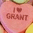 Pink heart-shaped candy with 'I ❤️ GRANT' text, held by a hand with yellow sleeve, background includes green and brown elements.