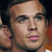 Pixelated close-up of a person's face with light skin tone, dark hair visible on the left side, and blurred background elements including brown and orange tones.