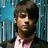 A person with short brown hair wears a pink shirt and dark jacket against a blue vertical striped background.