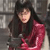 A pixelated figure with long dark hair wears a dark jacket over a red garment against a blurred background.