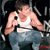 Pixelated image of a person in a white tank top and blue jeans holding a silver camera, standing against a dark background with metallic structures.