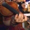 A person plays a violin with left hand on strings and right hand holding bow, while another person holds sheet music.