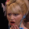 A close-up of a person with blonde hair styled in an updo, wearing a large blue and green beaded earring and a white feather accessory, with hands near the mouth.