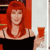 A person with long red hair wearing a black top holds a red glass against a red and white background.