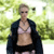 A person in a light pink bralette, black shorts, and open dark jacket walks outdoors with green foliage background.