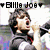 A person with dark tousled hair in a black and white jacket holds a microphone, mouth open as if singing or shouting, against a blurred background.