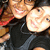 Two people in black hooded tops smile closely together, one with a red strap visible, against a blurred background.