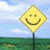 Yellow diamond-shaped sign with a smiley face on a pole, set against a blue sky and green field background.