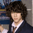Pixelated static image of a person with dark curly hair wearing a dark suit, white shirt, and striped tie against a blurred background.