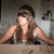 A pixelated image of a person with long brown hair wearing dark sunglasses and a sleeveless top, seated at a table in an indoor setting.