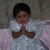 A child in a white dress with pink accents and angel wings lies on a light purple surface against a textured gray background, hands clasped in prayer.