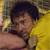 Pixelated image of a person with dark hair wearing a yellow shirt, holding a small object near their face against a blurred background.