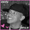 Black-and-white photo of a person wearing a cap and chain necklace, framed by pink border with 'sexy' text top-left and 'smile' text bottom-right, two pink hearts on left side.