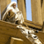 A pixelated static image shows a figure in white robes seated on a wooden structure with a dog-like animal beside them, set against a wooden wall and blue sky background.