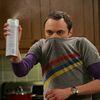A person in a gray shirt with purple sleeves and a rainbow stripe holds a white spray can emitting mist against a wooden background.