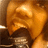 A pixelated close-up of a figure with light brown hair and a dark top, set against a warm-toned background with blurred horizontal elements.
