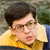 A pixelated close-up of a person with short dark hair wearing glasses and a yellow collared shirt against a blurred background.