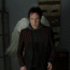 A person with large white angel wings stands in a dimly lit room with wooden crates in the background, wearing a dark jacket over a dark hoodie.