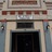 Static image of a building facade with a dark doorway framed by light-colored columns and decorative architectural details above.