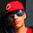 A person wearing a red cap with a white 'C' logo, dark sunglasses, and a red shirt, with a dark background.