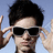 Pixelated image of a person with dark hair wearing black sunglasses, making a hand gesture near the face against a light blue background.