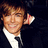 Pixelated image of a person in dark suit with white shirt and black tie, smiling while holding hand near ear, next to pink text reading 'Zac Efron is LOVEE<333'