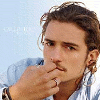 A person with light brown curly hair and a mustache wears a light blue striped shirt, hand near mouth in contemplative pose against light background.