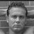Black-and-white mugshot-style image of a man with short hair against brick wall background, accompanied by three dark brown rectangular text boxes on white background.
