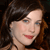 Pixelated close-up of a person with dark brown hair, light skin, blue eyes, and red lips, set against a dark background.