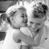 Two children in white clothing embrace closely, one with braided hair and the other with short hair, both smiling in a black-and-white photograph.
