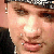 A pixelated close-up of a person's face with light skin tone, brown eyes, and a slight smile, wearing a dark jacket and a patterned cap.