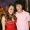Two people stand close together; the person on the left wears a red sleeveless dress with dark stripes, and the person on the right wears a light pink polo shirt.