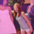 Pixelated image of a person with long brown hair wearing a light-colored top and dark sleeves, standing against a pink background.