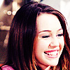 A close-up of a person with long brown hair smiling broadly, showing teeth, against a blurred background with warm tones.
