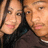 Two people in close-up, one with long dark hair and the other with short light hair, both wearing dark clothing, positioned side-by-side against a light background.