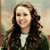 A person with long curly brown hair wearing a green patterned jacket over a white shirt and a beaded necklace, smiling with eyes partially closed.