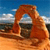 Pixelated landscape showing a red rock arch structure against blue sky with clouds and distant terrain.