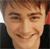 A pixelated close-up of a smiling person with light brown hair, wearing a dark top against a blurred background with red and white elements.