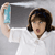 A person with brown hair in a white shirt holds a light blue cylindrical object against a gray background.