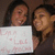 Two people with dark hair hold a white sign with pink text against a brown background.