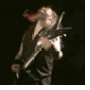 A person with long flowing hair plays a black electric guitar against a dark background, captured in showing motion