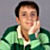 Pixelated static image of a person with short dark hair wearing a green and white collared shirt, hand near face, against gray background.
