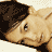 Pixelated image of a person lying down in a light-colored outfit with visible tattoos on the arm and chest, set against a light background with repeated text watermark.