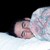 Pixelated static image of a person lying down with eyes closed, resting on a light surface against dark background.