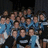 A static pixelated image shows a group of people in blue and white clothing against a dark background with no visible text or distinct symbols.