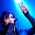 A person with dark hair sings into a microphone under blue stage lighting, making a peace sign gesture while red text reads "the best part of 'believe' is the 'lie'" on black background.