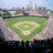 A baseball field with green grass and brown dirt, surrounded by stadium seating under a blue sky.