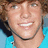Pixelated close-up of a smiling person with light skin, brown hair, and visible teeth against a blue background.