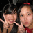 Two people with dark hair pose closely together against a dark background, one wearing a red headband and red clothing, the other with a hand near their face.