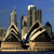 Pixelated cityscape with tall buildings under blue sky, featuring a prominent tower with a spire and a larger structure with rounded top.